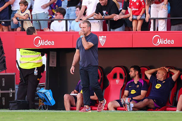 Hansi Flick stands on a soccer field near a bench with red seats, wearing a dark shirt and red shoes. Players in FC Barcelona uniforms sit on the bench, some resting their heads on their hands. A security guard in a yellow vest and spectators in the stands are visible in the background. Midea logos are displayed on the bench area. Hansi Flick stands on a soccer field near a bench with red seats, wearing a dark shirt and red shoes. Players in FC Barcelona uniforms sit on the bench, some resting their heads on their hands. A security guard in a yellow vest and spectators in the stands are visible in the background. Midea logos are displayed on the bench area.