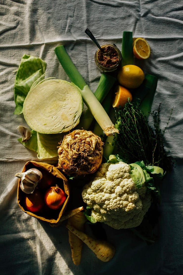 An overhead shot of winter vegetables on a white linen background in harsh light. There is: a halved green cabbage, a head of cauliflower, a bag of onions and garlic, some parsnips, a celery root, lemons, and leeks.