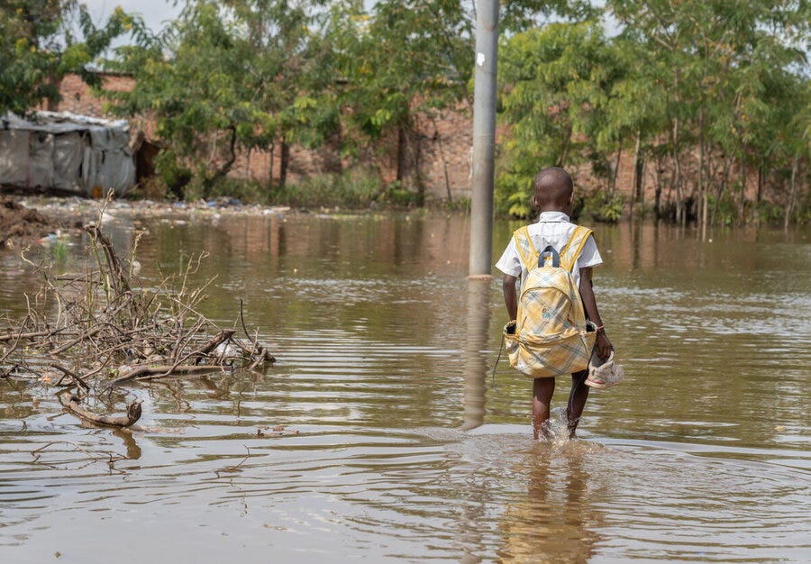 Child with backpack walks through knee‑deep floodwater, holding shoes, as homes, trees and debris line a flooded street in a damaged neighbourhood. Child with backpack walks through knee‑deep floodwater, holding shoes, as homes, trees and debris line a flooded street in a damaged neighbourhood.