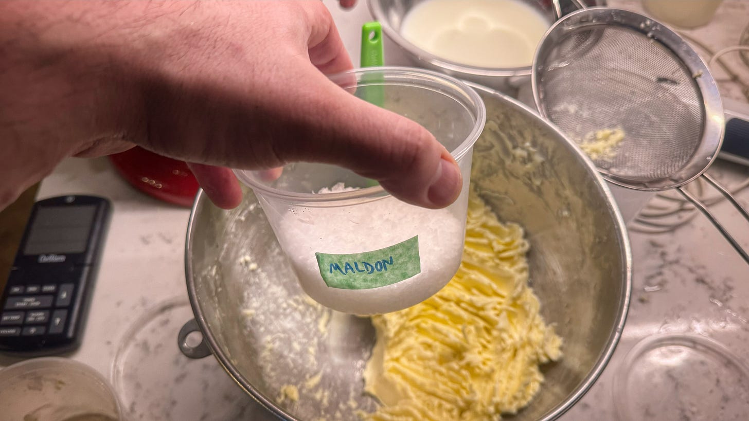 A hand holding a deli container full of flaky salt marked "Maldon." Behind it we can see the mixing bowl with the completed butter. A hand holding a deli container full of flaky salt marked "Maldon." Behind it we can see the mixing bowl with the completed butter.