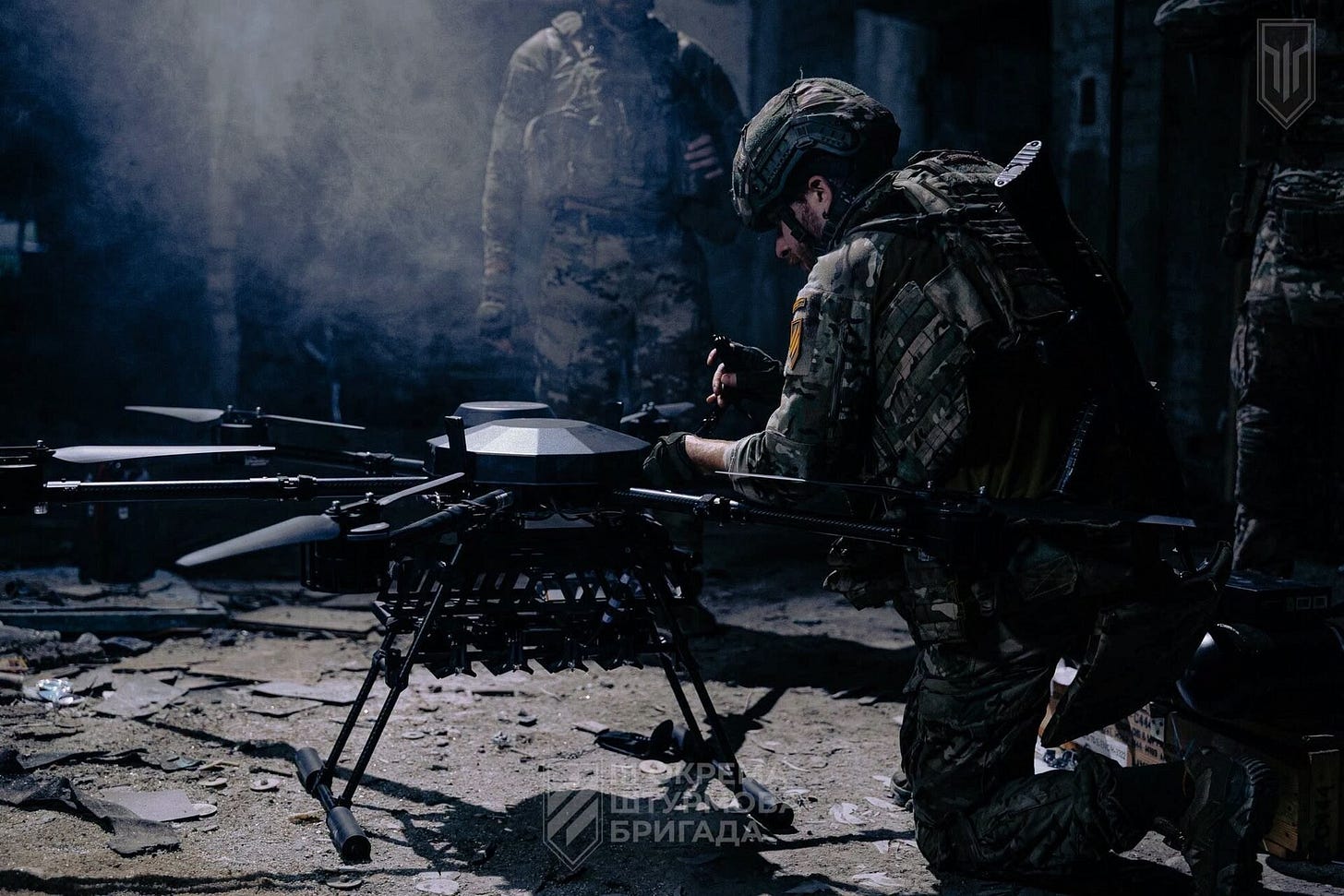 A Ukrainian soldier in a helmet and body armor kneels to prepare a large multi-rotor bomber drone inside a smoky, damaged building while another soldier stands behind him. A Ukrainian soldier in a helmet and body armor kneels to prepare a large multi-rotor bomber drone inside a smoky, damaged building while another soldier stands behind him.
