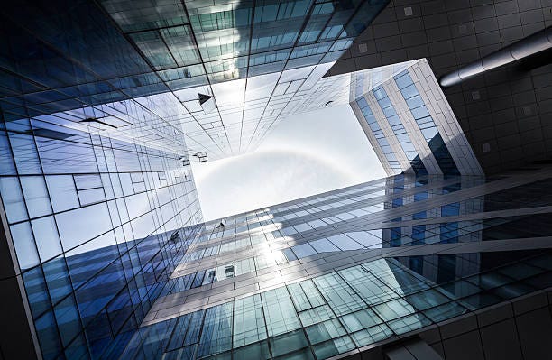 Looking up at the modern glass architecture of a building, showcasing sharp lines, reflective surfaces, and geometric patterns against a bright sky.
