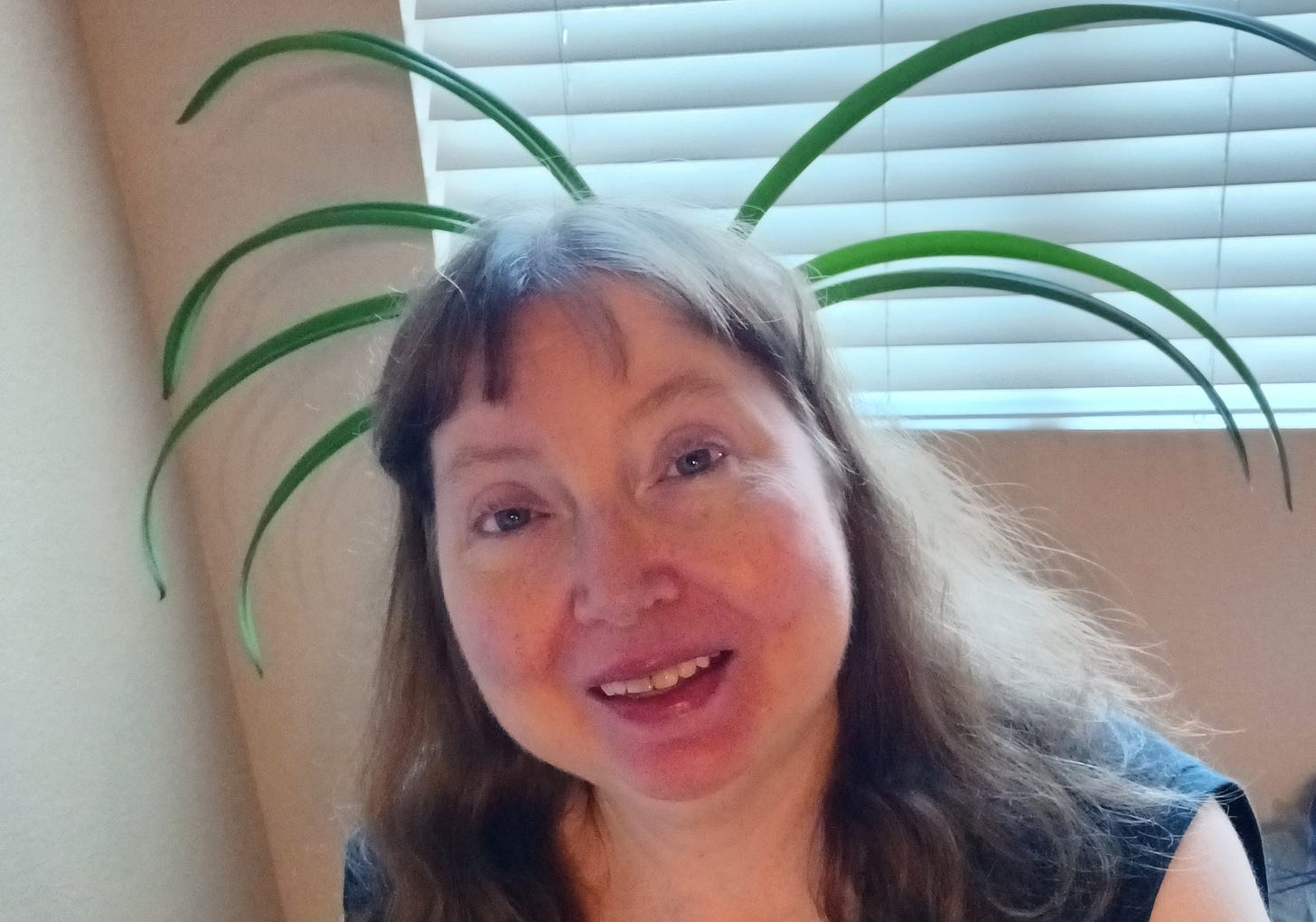 a woman with auburn hair and blue eyes, wearing a dark shirt, has multiple giant green plant leaves sprouting in all directions behind her head, and behind it all are sunlit white window blinds