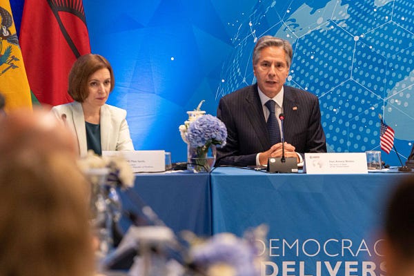 Secretary of State Antony Blinken sits at a rectangular table with blue table cloth that says "Democracy Delivers". He is speaking into the microphone during a USAID-Organized Democracy Delivers Event at the UNGA.