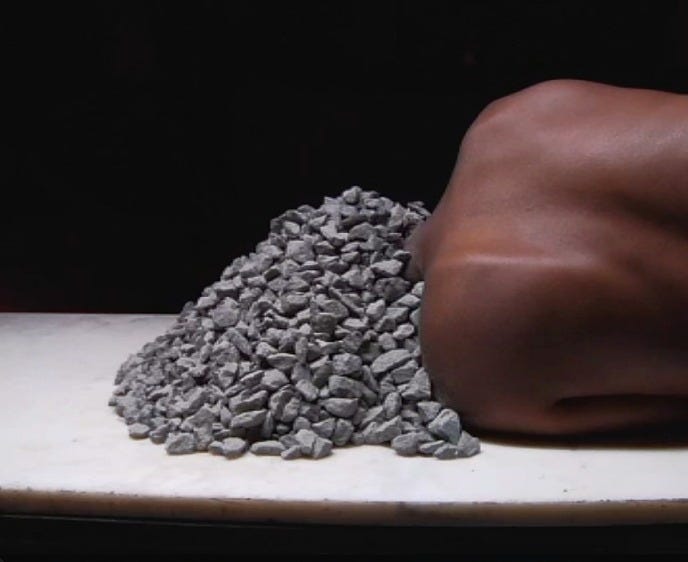 A black woman’s naked back as she lays on a white surface. She has several small gray rocks that seem to have fallen on her and covered her entire head. 