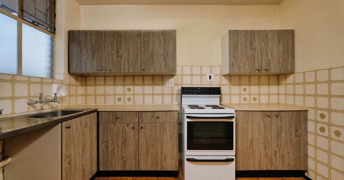 A dated kitchen with gray wood-grain cabinets, a white electric range and cream wall tiles with a gold flower pattern