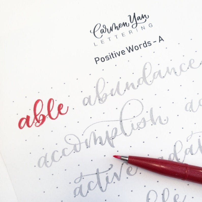 May include: A close-up of a sheet of paper with gray dots. The word "able" is written in red ink. The words "abundance", "accomplish", and "active" are written in gray ink. A red pen is lying on the paper.