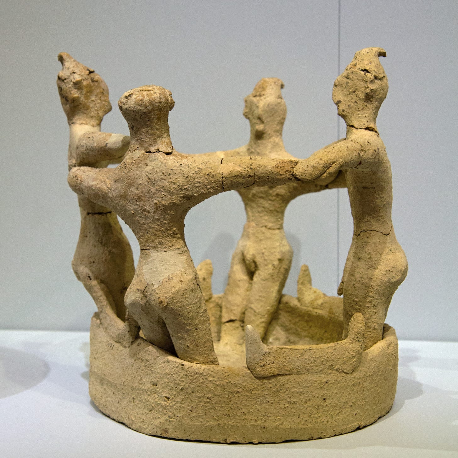 Rough terracotta figurine showing four human figures of indistinct gender wearing loincloths and pointed hats. They're all standing in a circle in a large vat that comes up to their knees, with their arms interlinked. The sides of the vat are decorated with Minoan sacred horns.