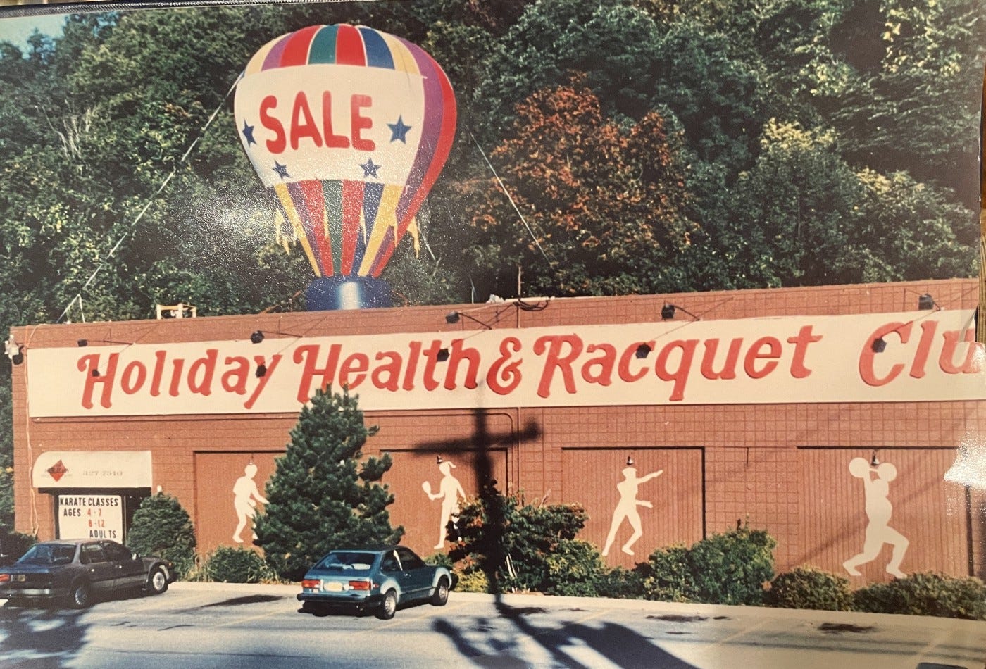 A true-life Kodachrome filter, a parking lot fronting a brick building topped by the words “holiday health and racquet club”