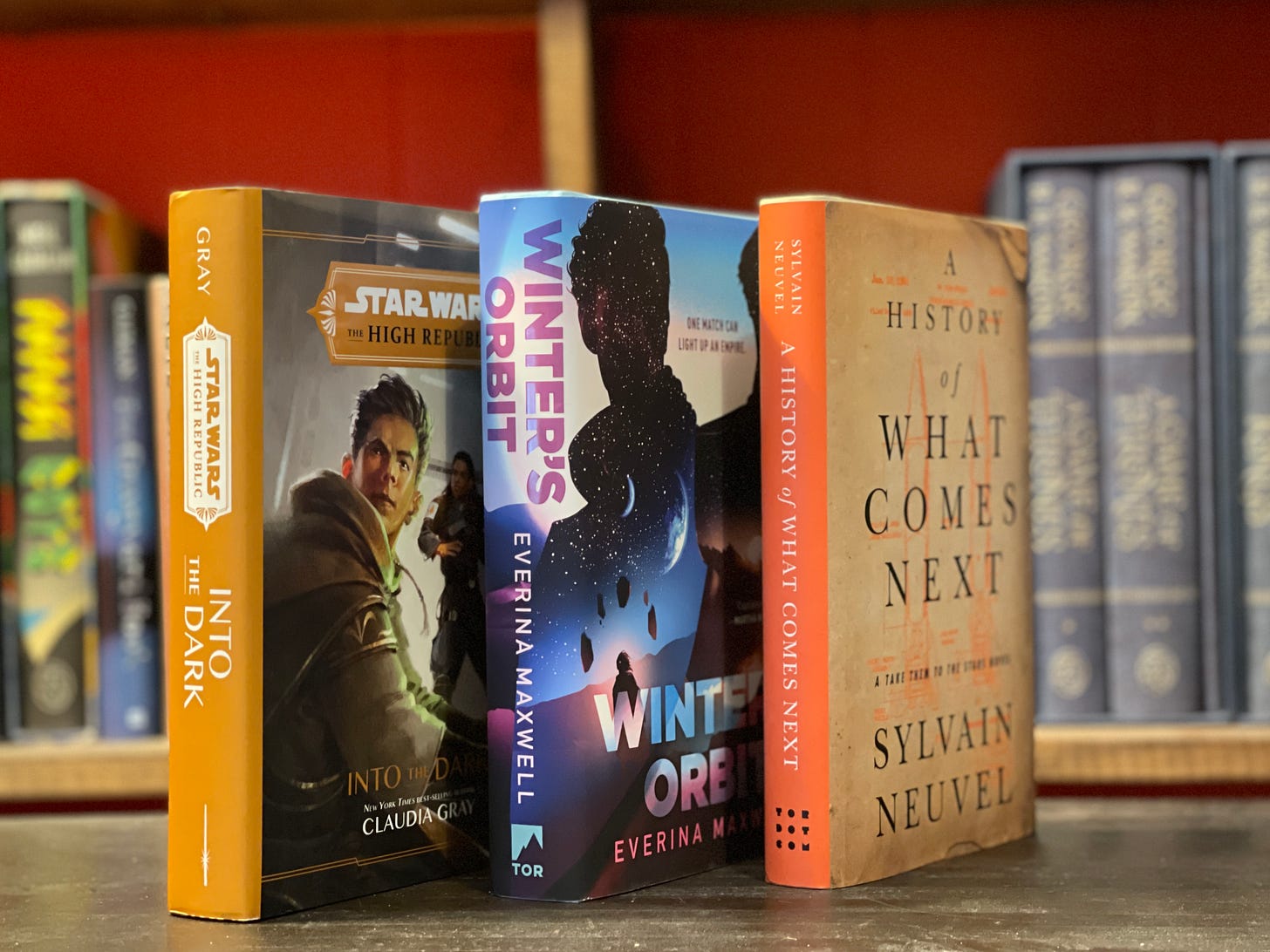 Three books standing on a table: Into the Dark, Winter's Orbit, and History of What Comes Next