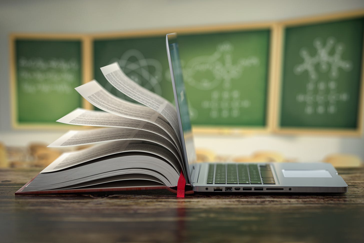 Desk with an open laptop and a textbook leaning against it. Blurred chalkboards in the background.