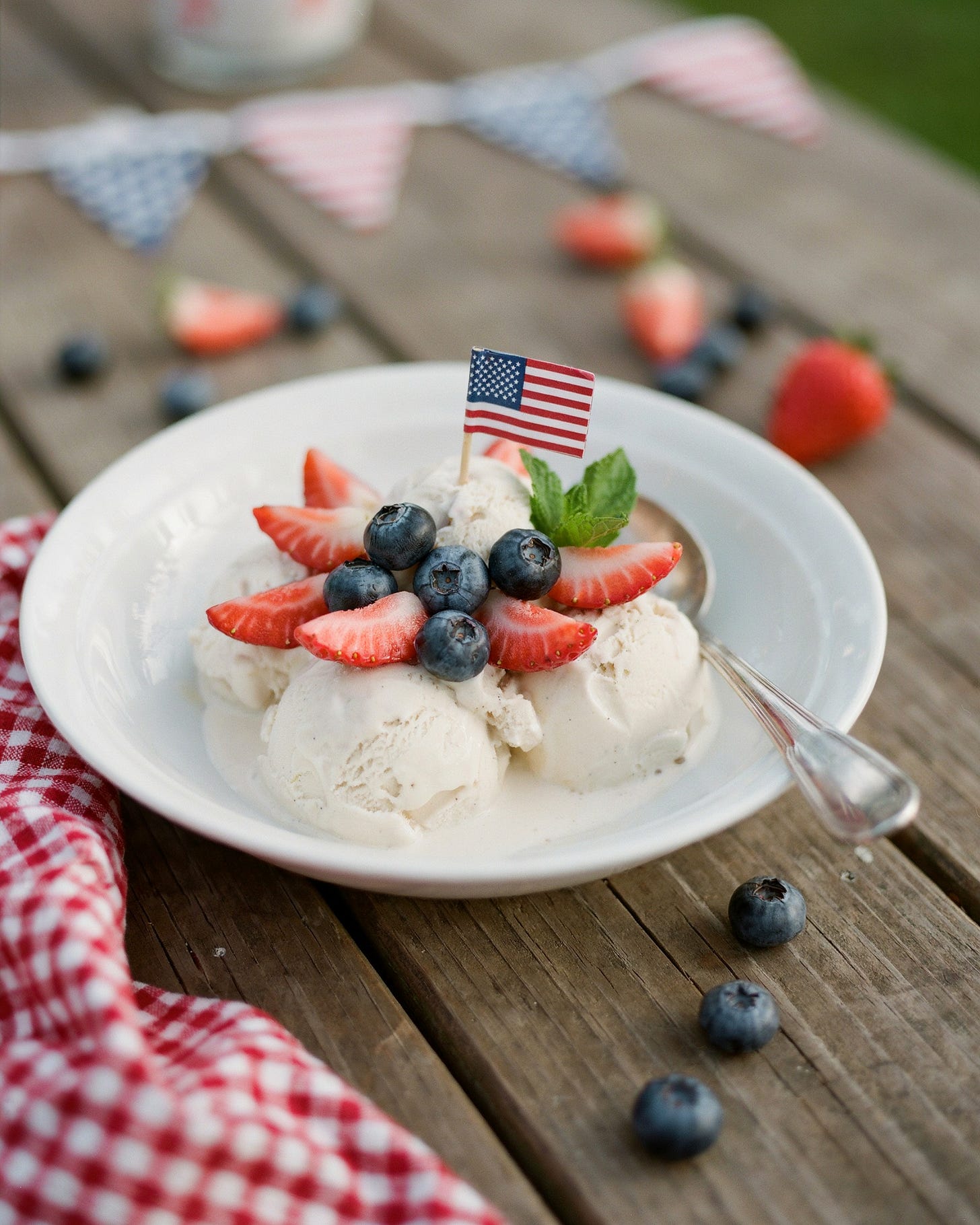 a white bowl full of vanilla ice cream, berries and an American flag on top of a picnic table