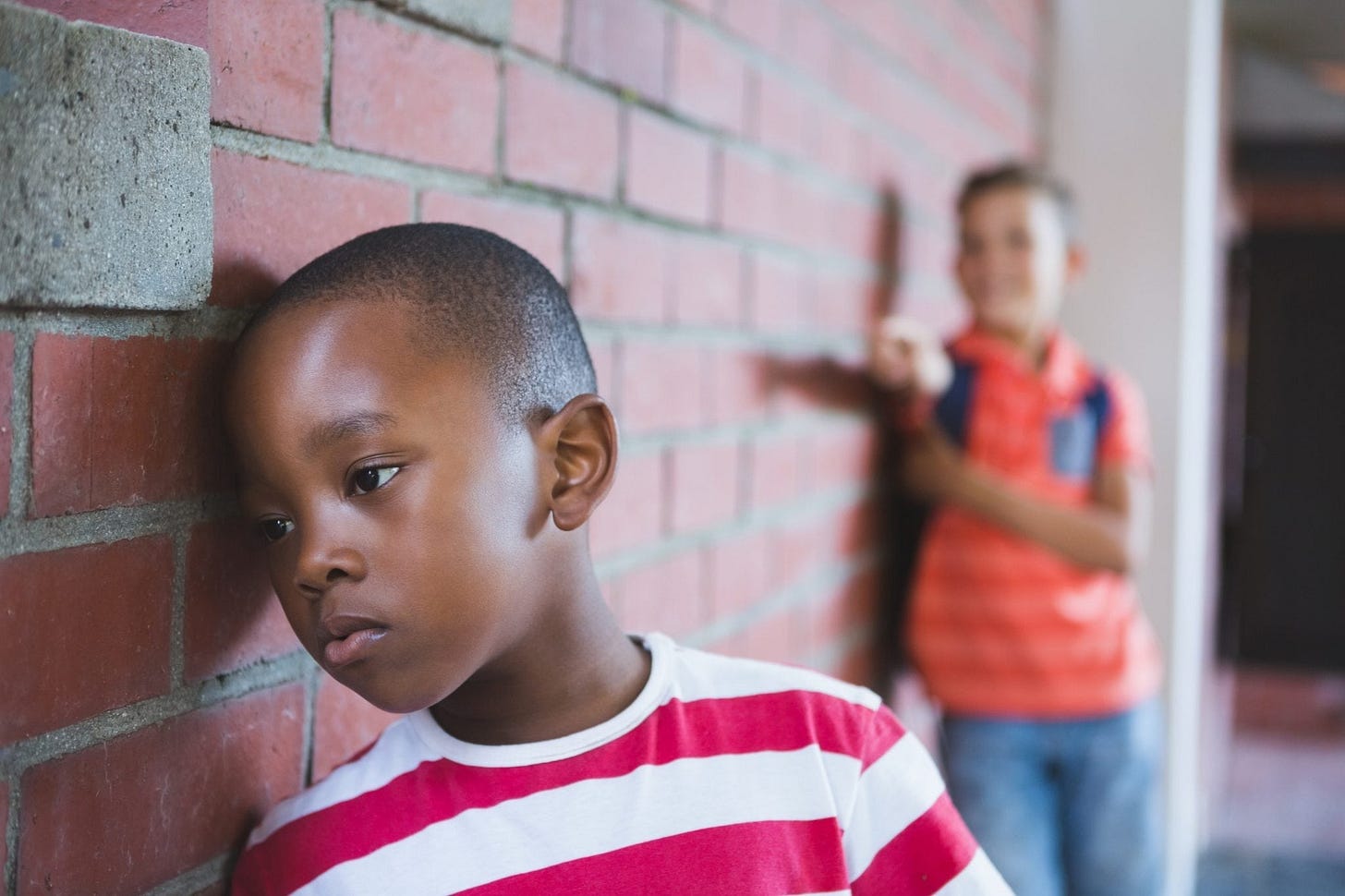 Een jongen in een rood-wit gestreept shirt leunt verdrietig tegen een bakstenen muur, terwijl een andere jongen op de achtergrond toekijkt met een ernstige blik. Een jongen in een rood-wit gestreept shirt leunt verdrietig tegen een bakstenen muur, terwijl een andere jongen op de achtergrond toekijkt met een ernstige blik.