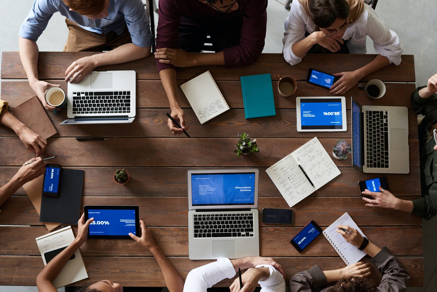 A photograph taken from overhead of a wooden table, on which we see three laptops, several tablets, several notebooks, and the hands of eight people gathered around that table