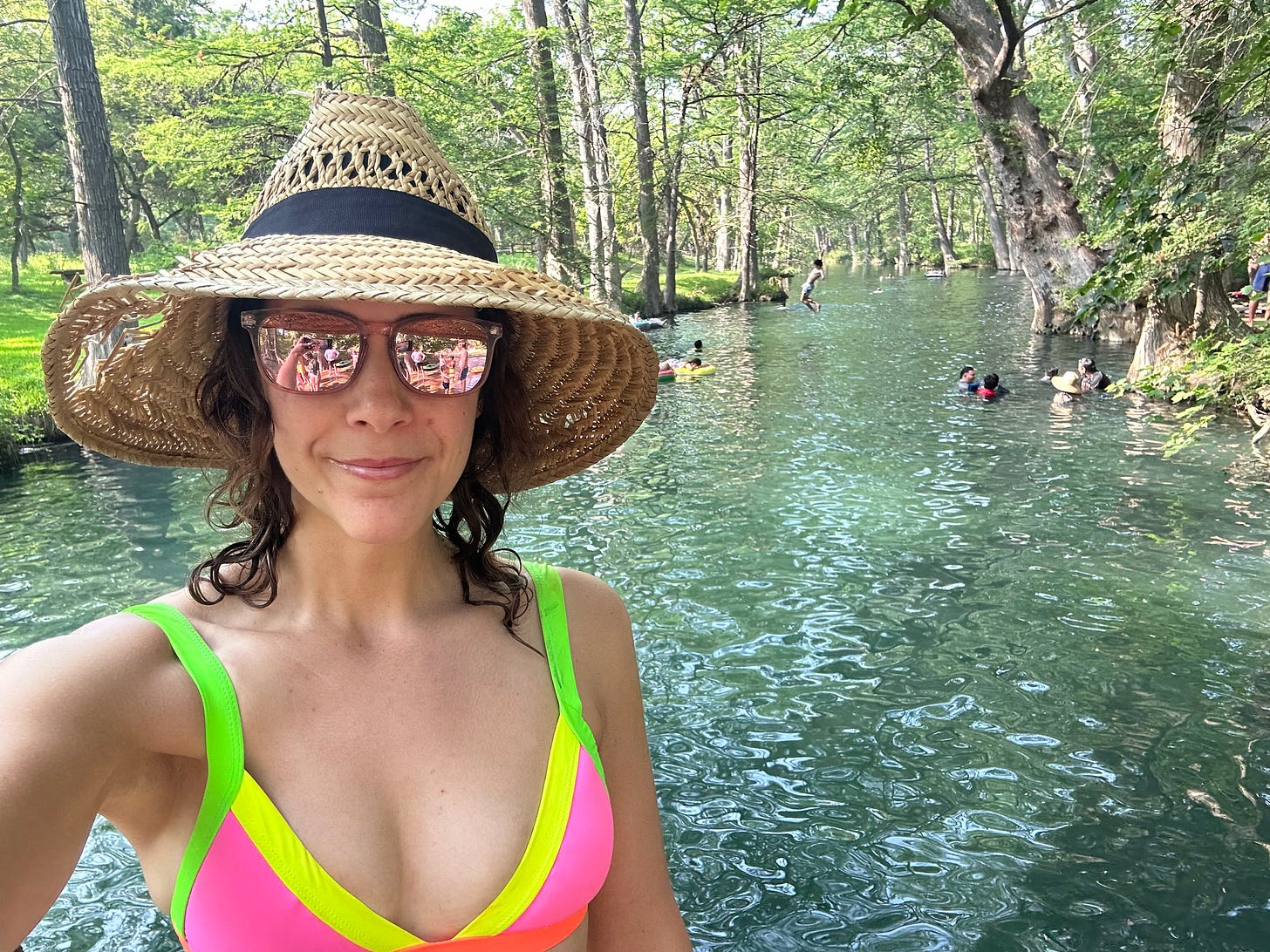 A women wearing a pink bikini top, reflective sunglasses, and a straw hat takes a selfie with a swimming hole behind her. A women wearing a pink bikini top, reflective sunglasses, and a straw hat takes a selfie with a swimming hole behind her.