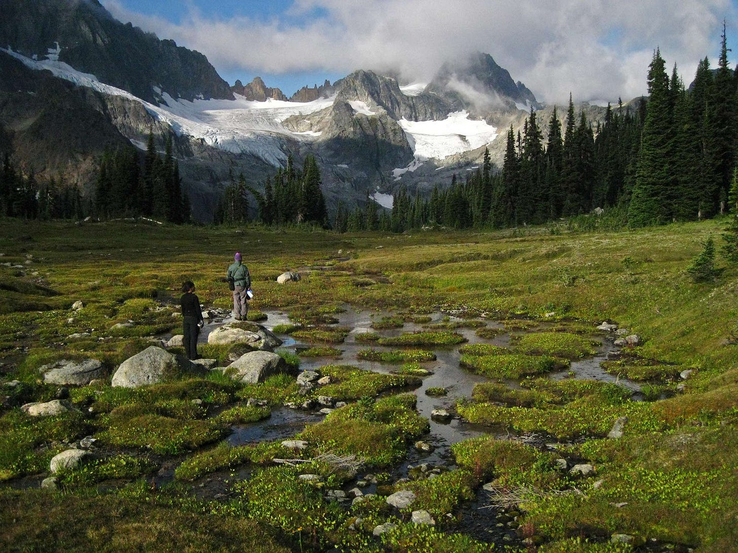 a man and woman look up at a sunlit mountain peak veiled with clouds from a bright green meadow and alpine creek dotted with boulders and tiny green mossy islands