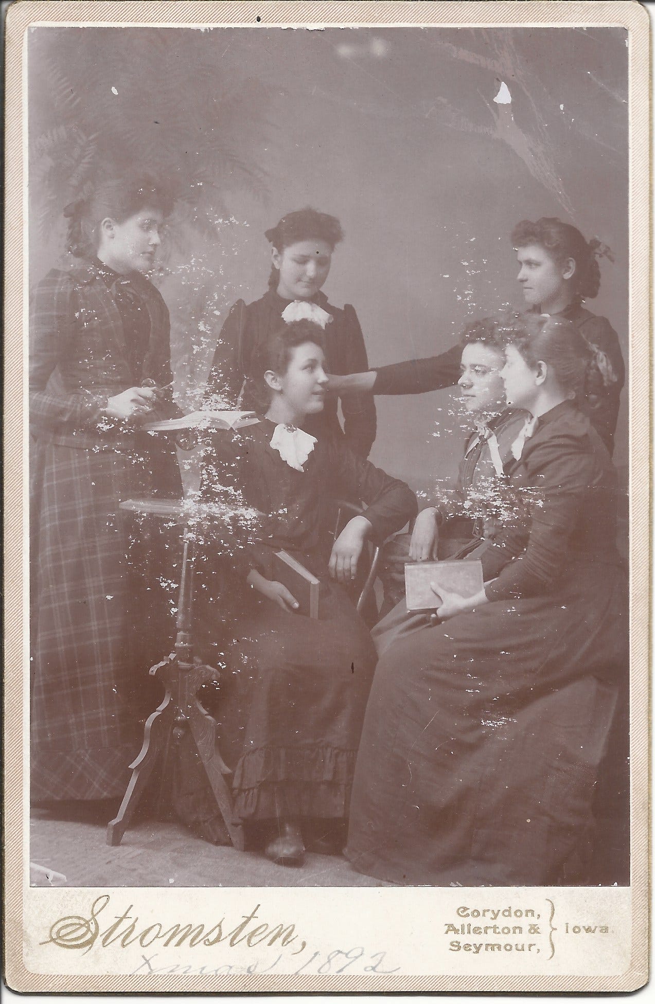 Black and white photo of six young white women in Victorian dress, gathered around a small table. The woman on the left is Erna Ammons Gordon and the photo is labeled "Cousins and Friends" on the back. C. 1895