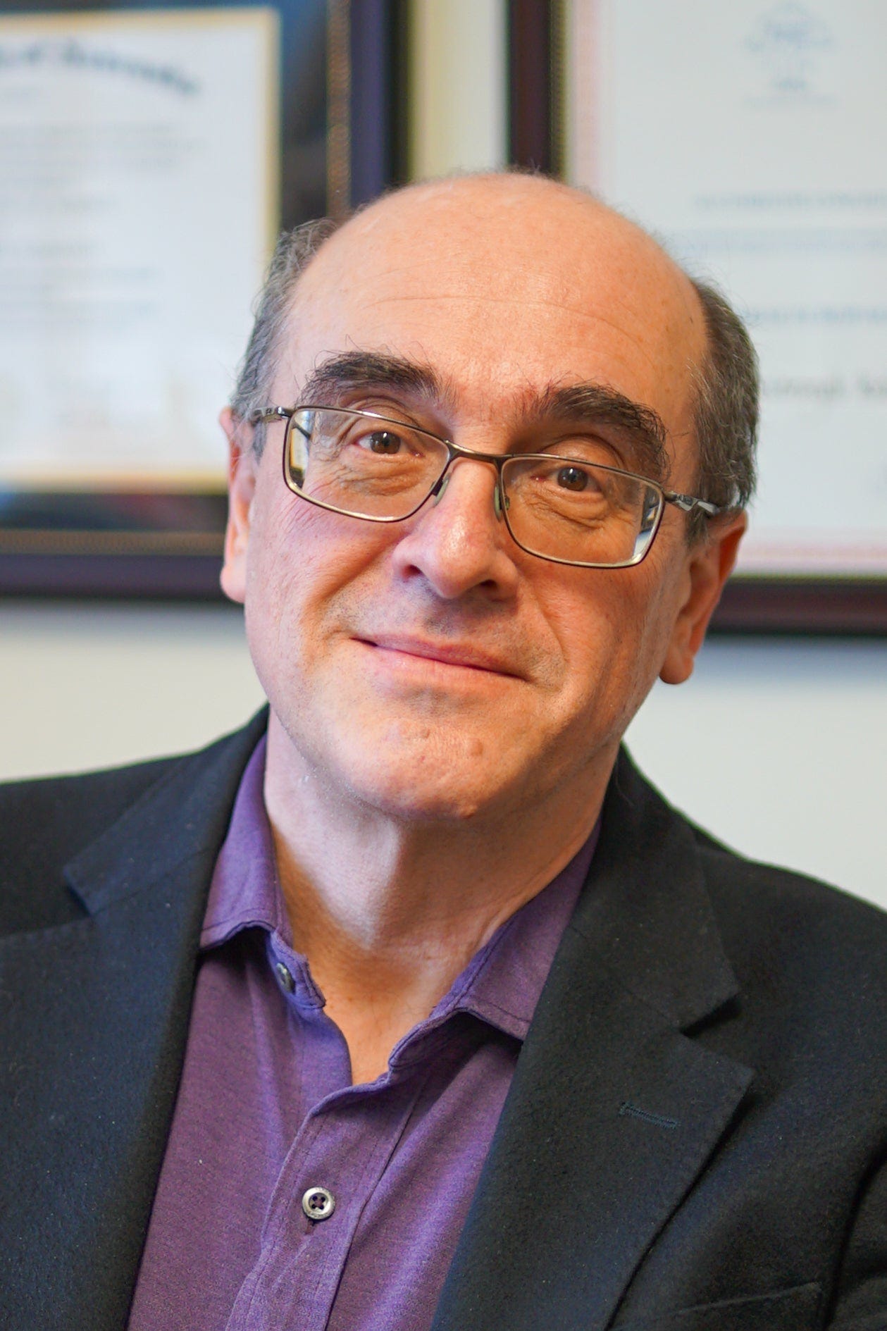 Sasha Kabanov in a purple shirt and black coat smiles in his office