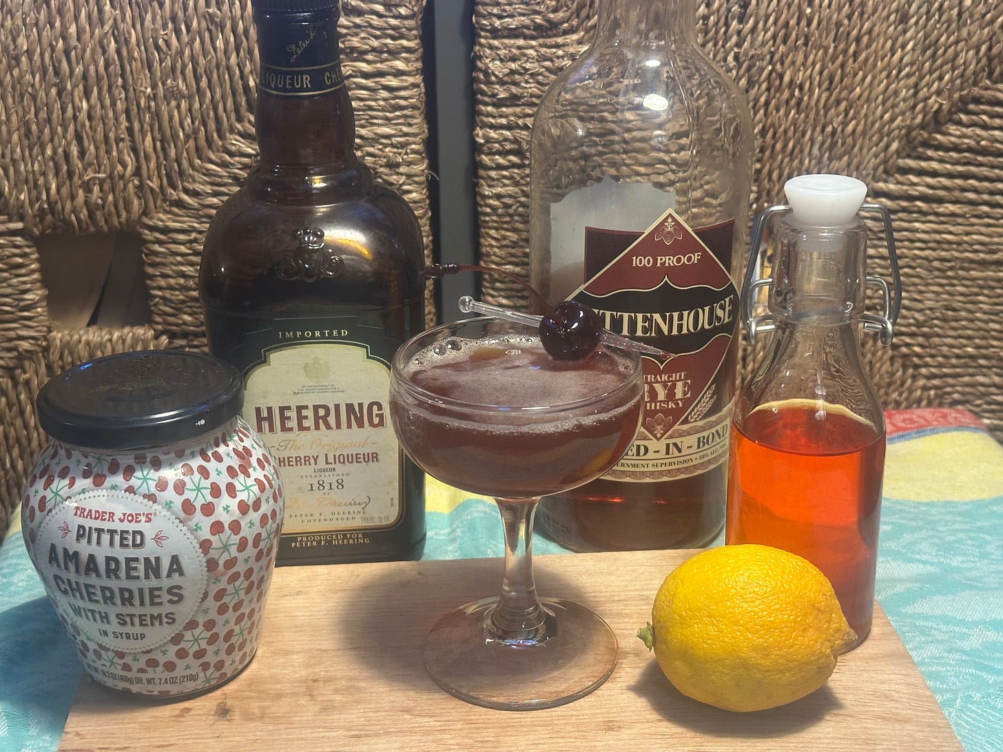 Photo of the ingredients (jar of pitted amarena cherries, Heering cherry liqueur, bottle of Rittenhouse rye whiskey, orangish infusion of chili pepper in Aperol, and a lemon, arranged around the titular cocktail. 
