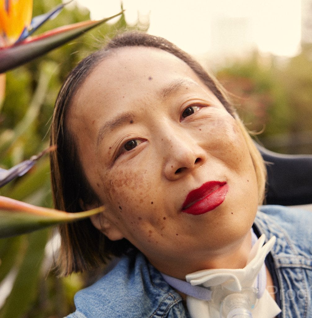 Alice Wong, an Asian American disabled woman with a tracheostomy at her neck. She is wearing a bright red lip color and a denim shirt. She is smiling with her head tilted toward some bird of paradise plants.