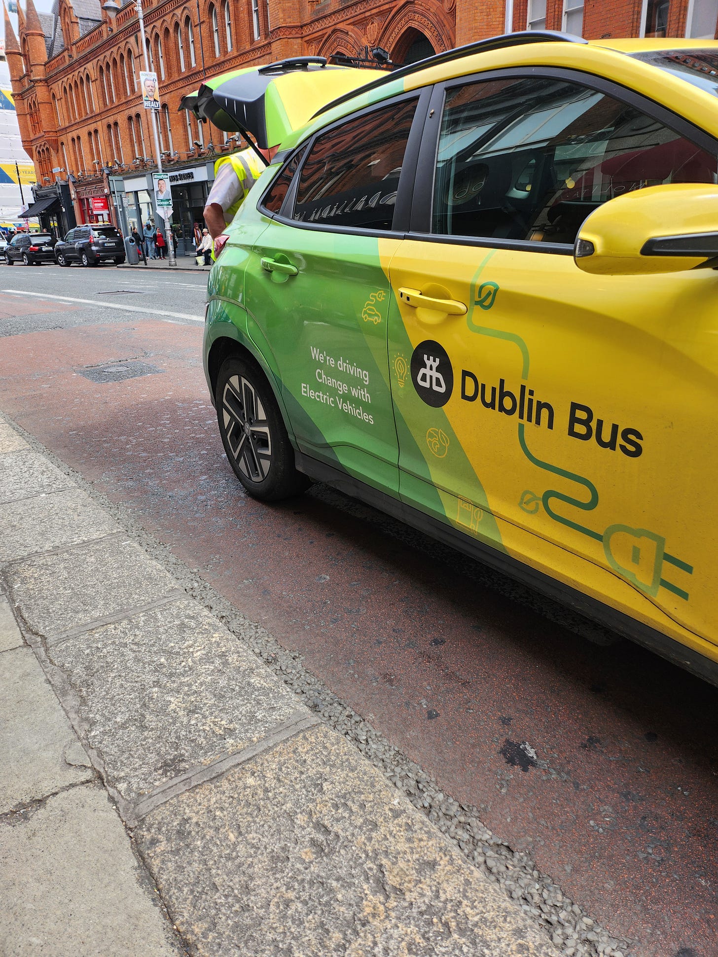 A yellow and green car with the Dublin Bus logo and the caption "we're driving change with electric vehicles" is pulled into the kerb at the side of George's St. A man in a hi-vis is rummaging in the boot. 