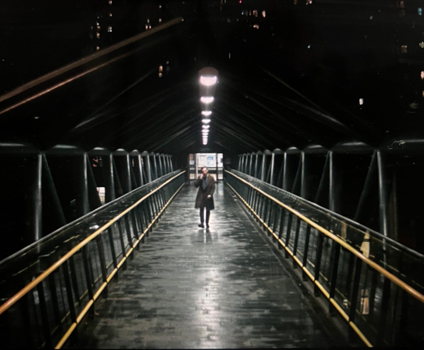 A person walks down a long, covered pedestrian bridge at night. The walkway is empty, slick with rain, and lit by evenly spaced overhead lights. The bridge's glass sides and metal beams frame the figure at the center.