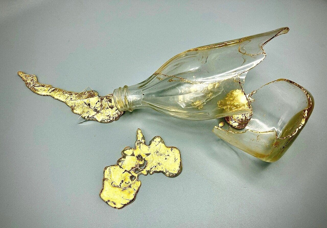 A glass bottle broken in two pieces lies on a grey surface. Cracks in each of the two pieces are lined with gold, and two small puddles of gold seem to spill out next to the bottle on the surrounding surface.