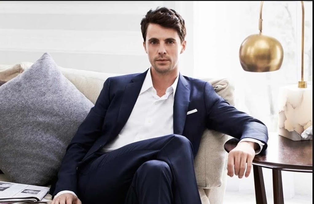 Medium shot of a tall, slender brown-haired man wearing a blue suit sitting on a sofa. 