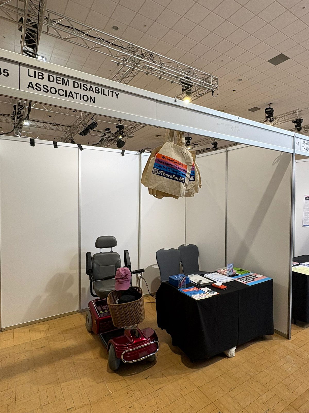 A conference booth with a sign reading “Lib Dem Disability Association.” The booth has plain white partition walls, a table covered with a black cloth displaying flyers, booklets, and a few items, and two empty chairs behind it. In front, there is a red mobility scooter with a basket containing a pink hat. Hanging from the booth frame is a beige tote bag printed with a campaign message about NHS care for people with ME and Long Covid.