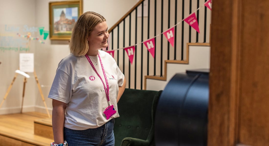 A woman wearing a white t-shirt and a pink lanyard stood in front of steps with pink bunting on A woman wearing a white t-shirt and a pink lanyard stood in front of steps with pink bunting on