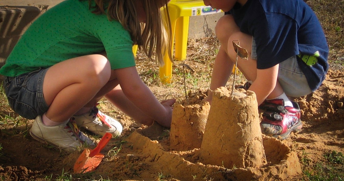 a young girl in a green shirt and jean shorts with a young boy in a blue shirt and light blue shorts squating to build sandcastles