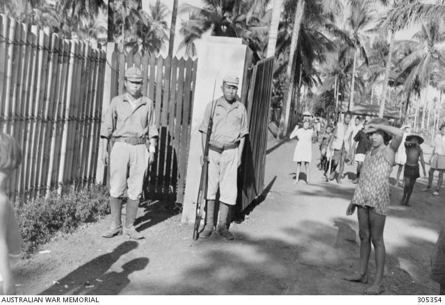 Two Japanese camp guards and a civilian girl look towards the photographer capturing the scene outside the gate of the prison after the end of the Second World War. Two Japanese camp guards and a civilian girl look towards the photographer capturing the scene outside the gate of the prison after the end of the Second World War.