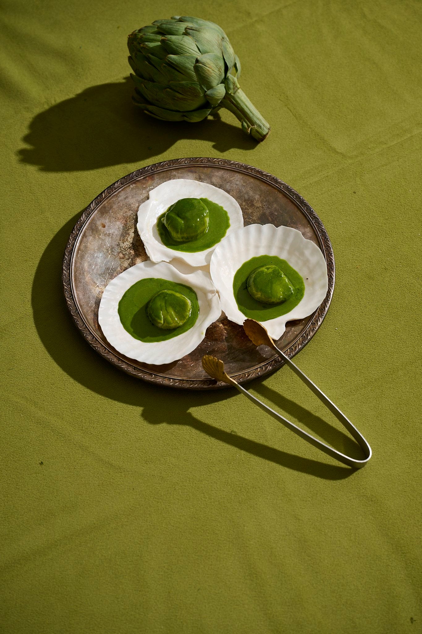 Scallops in the shell with vibrant green herb butter, served on a silver tray with vintage tongs, set against a green tablecloth with a fresh artichoke.