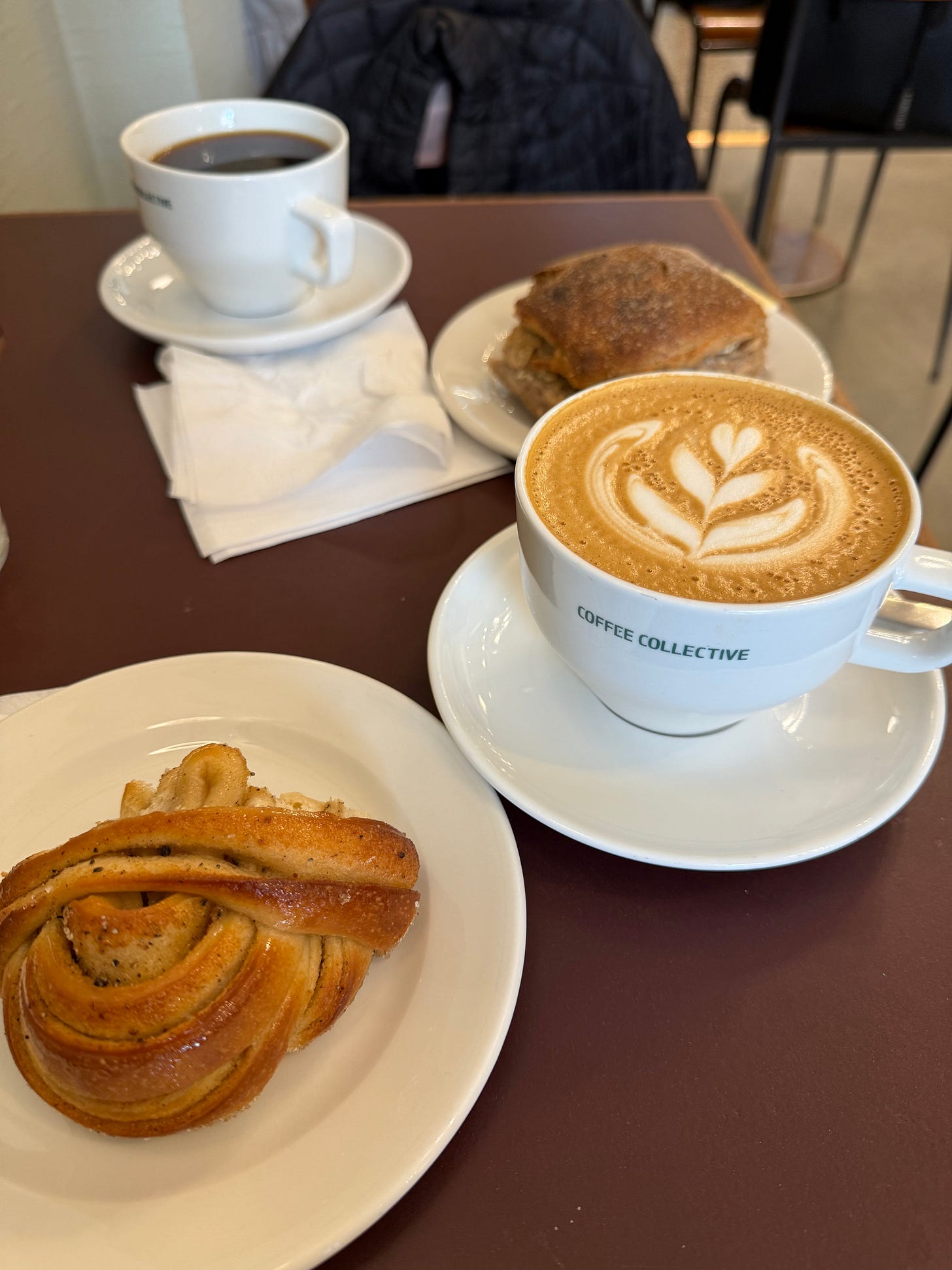 A cozy café table at Coffee Collective Bernikow in Copenhagen, featuring a cappuccino with elegant latte art, a black coffee, a cardamom bun, and a sandwich on rustic bread. A cozy café table at Coffee Collective Bernikow in Copenhagen, featuring a cappuccino with elegant latte art, a black coffee, a cardamom bun, and a sandwich on rustic bread.