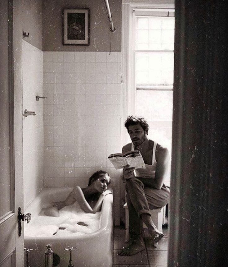 This may contain: a man sitting in a bathtub next to a woman laying on the floor and reading