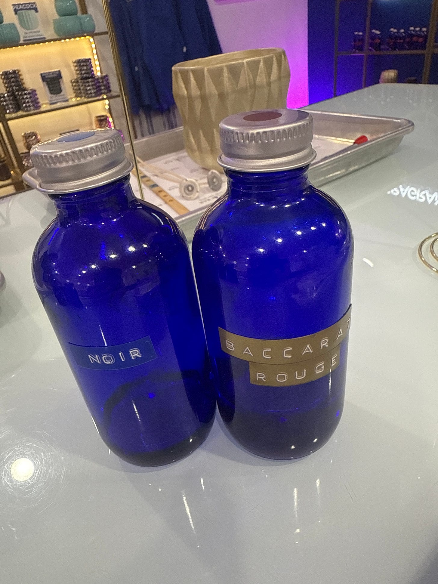 Two blue glass bottles with label maker labels reading 'Noir' and 'Baccarat Rouge' on a white counter Two blue glass bottles with label maker labels reading 'Noir' and 'Baccarat Rouge' on a white counter