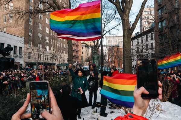 A Pride flag flies over a small city park surrounded by onlookers. A woman raises her fist in the air underneath the flagpole.