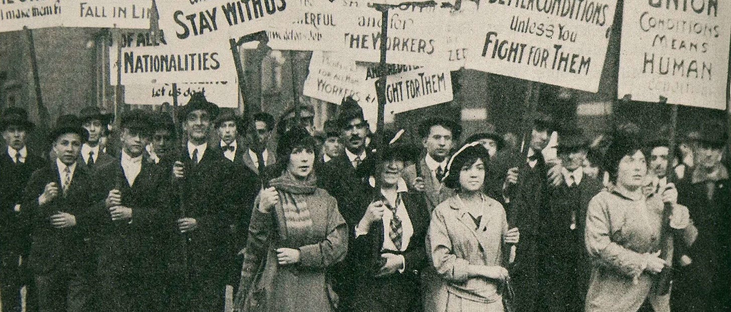 Historical black and white photograph of a workers' protest and rally. A crowd of people, both men in suits and women in long coats, stand together holding signs with visible text including 'Stay With Us', 'Fall In Line', 'Fight For Them'. The photograph is from the early 20th century.