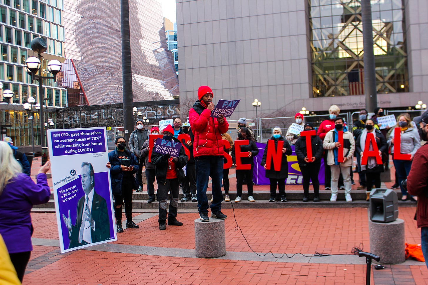 a line of people holding big red letters spelling "ESSENTIAL" stands behind a black man in a red coat and hat holding a "pay essential workers" sign a line of people holding big red letters spelling "ESSENTIAL" stands behind a black man in a red coat and hat holding a "pay essential workers" sign