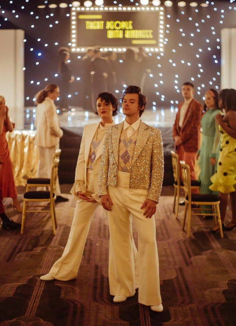 A colour behind the scenes shot of the Treat People with Kindness Music video. Harry Styles and Phoebe Waller-Bridge stand in the foreground looking at the camera with ambiguous expressions. They are both wearing the sparkly suits from the music video. Behind them is the set of the music video and the extras.