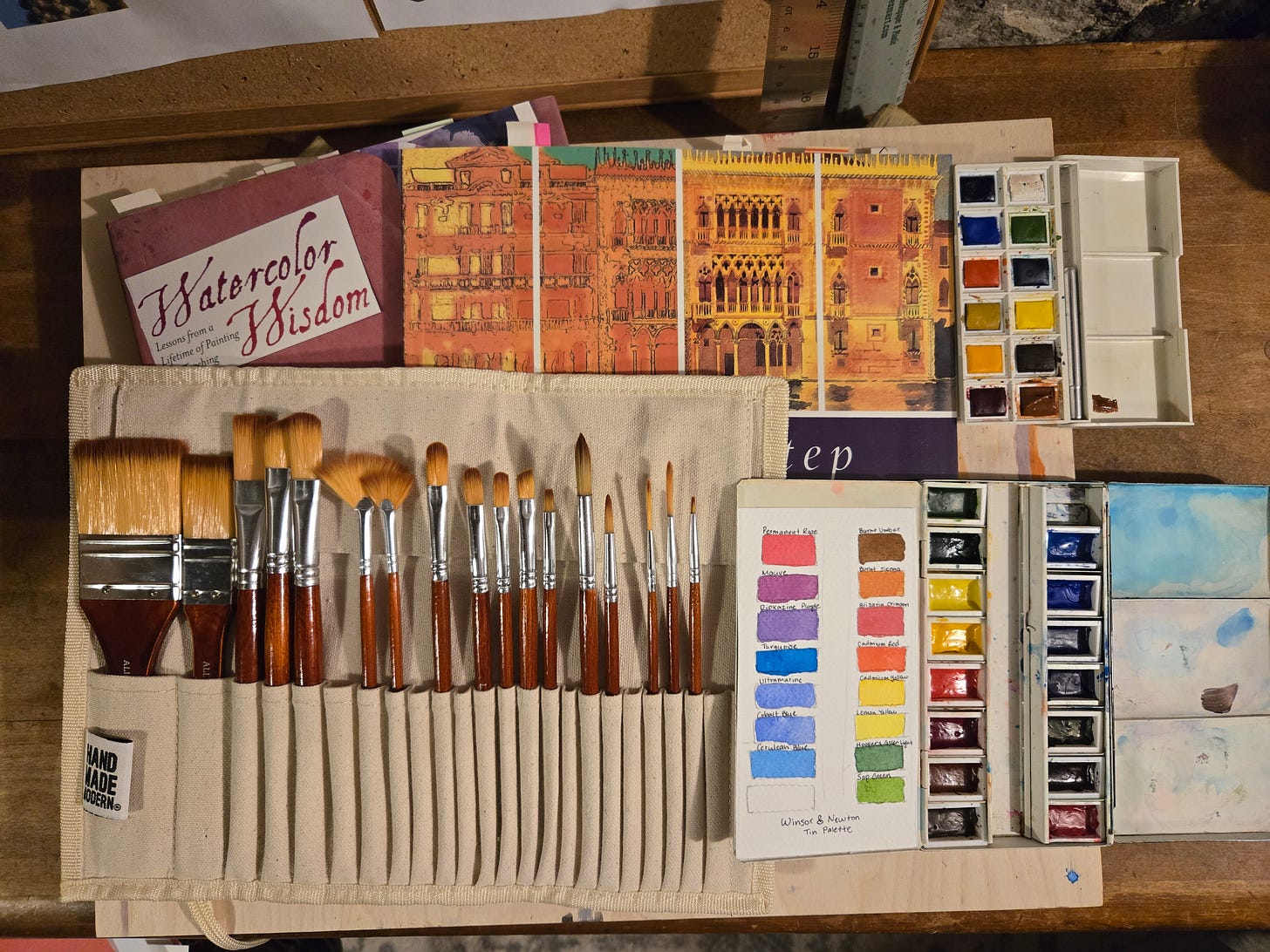 Flat lay of watercolor painting supplies including a set of paintbrushes in a canvas holder, Winsor & Newton watercolor palettes, a color swatch chart, sketchbooks, and the book Watercolor Wisdom on a wooden desk.