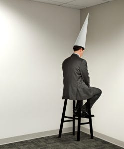 A man in a business suit sitting on a stool facing the corner of a room wearing a tall triangular hat