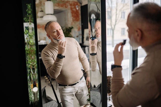 A middle aged white man with a gray beard, neatly trimmed, wearing a being turtleneck and light beige pants, combs his beard in a mirror.