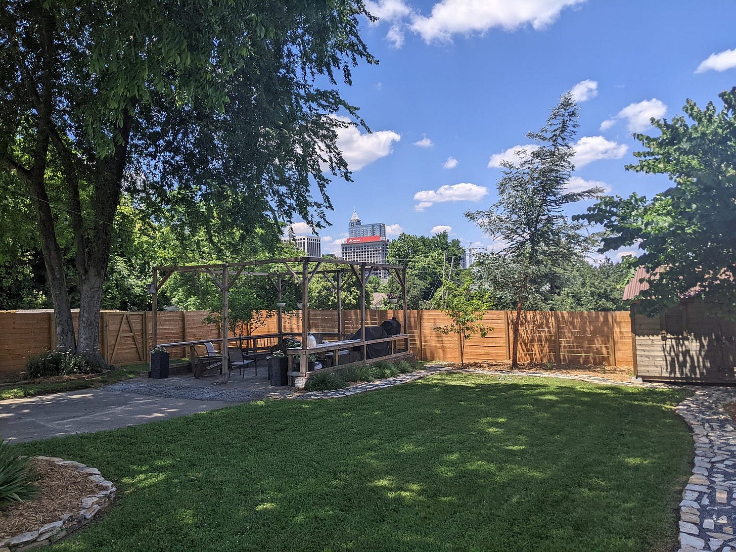 A backyard with green grass, a pergola, and a fenced-in yard with a view of the city in the background, with blue sky and puffy clouds. A backyard with green grass, a pergola, and a fenced-in yard with a view of the city in the background, with blue sky and puffy clouds.