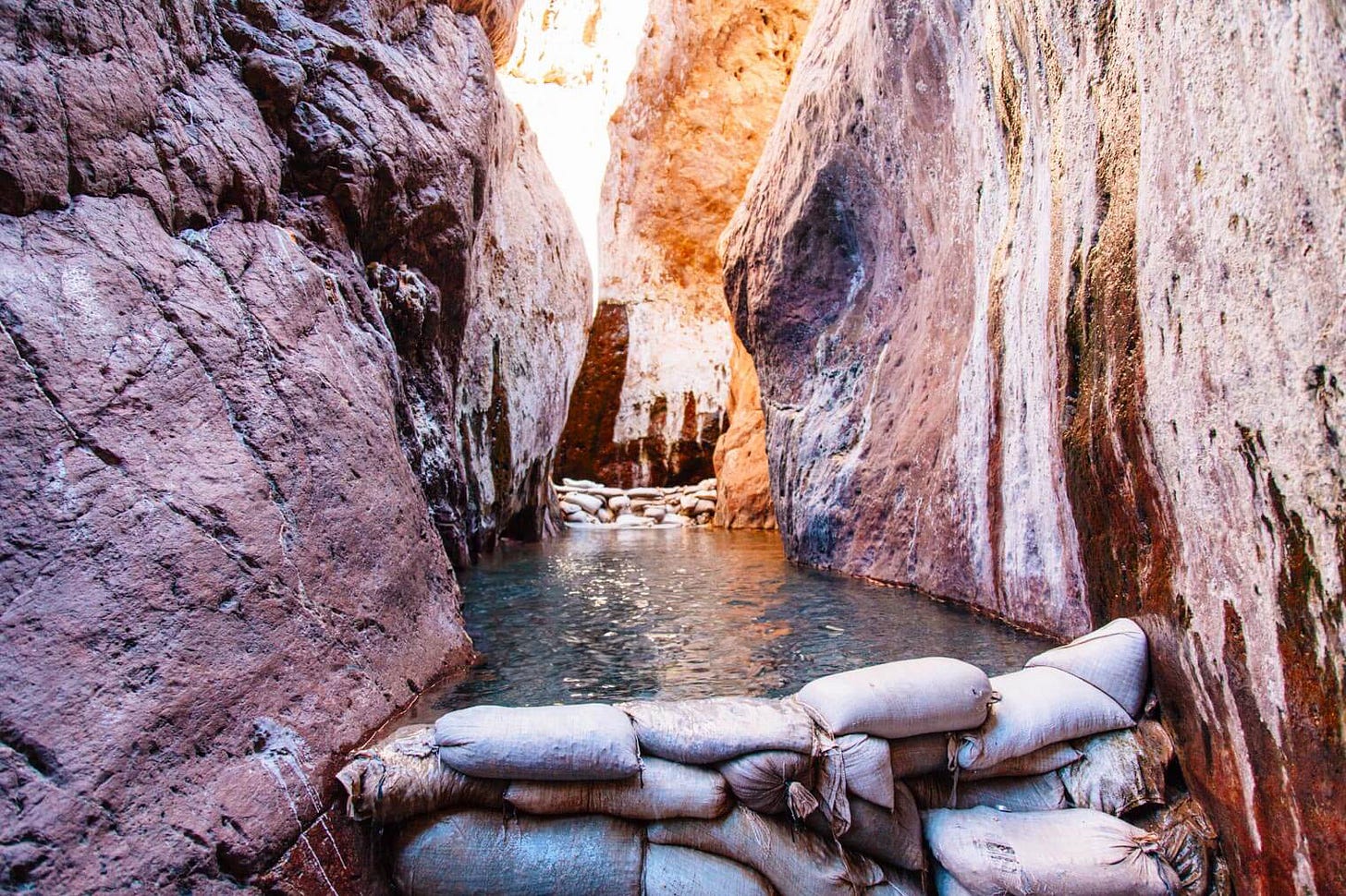 View upstream through a narrow slot canyon at Arizona Hot Springs. Warm water fills a series of pools separated by stacked sandbag dams, with mineral-stained canyon walls rising steeply on both sides.