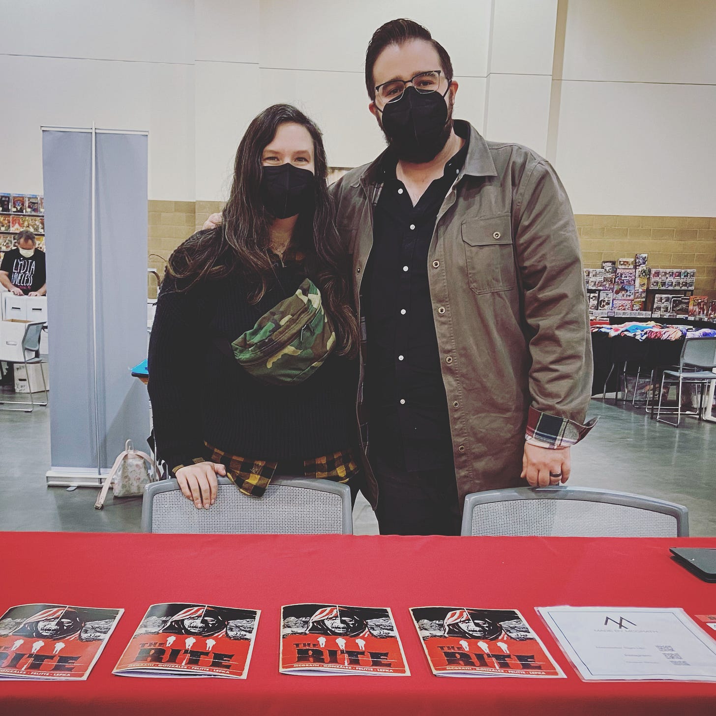 Two people wearing masks stand behind a table with a red cloths and printed books of The Rite.