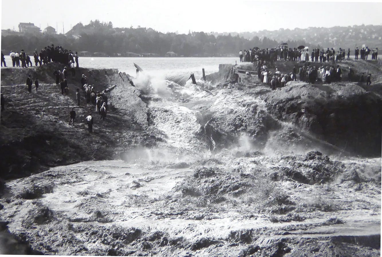 People stand on each side of a torrent of water coming through an earthen wall