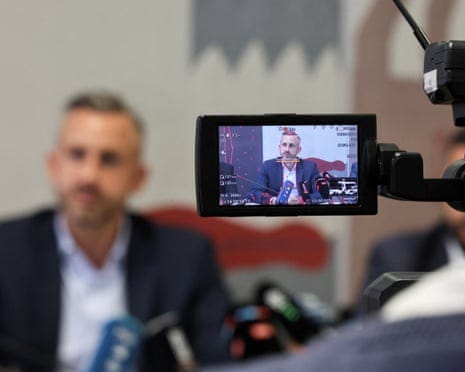 Camera screen shows Ulrich Siegmund speaking at microphones during a press conference