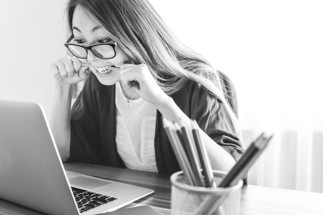 woman biting pencil while sitting on chair in front of computer during daytime