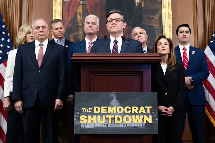 A group of people standing behind a man who's standing at a lectern, behind a sign that says 'The DEMOCRAT SHUTDOWN.'